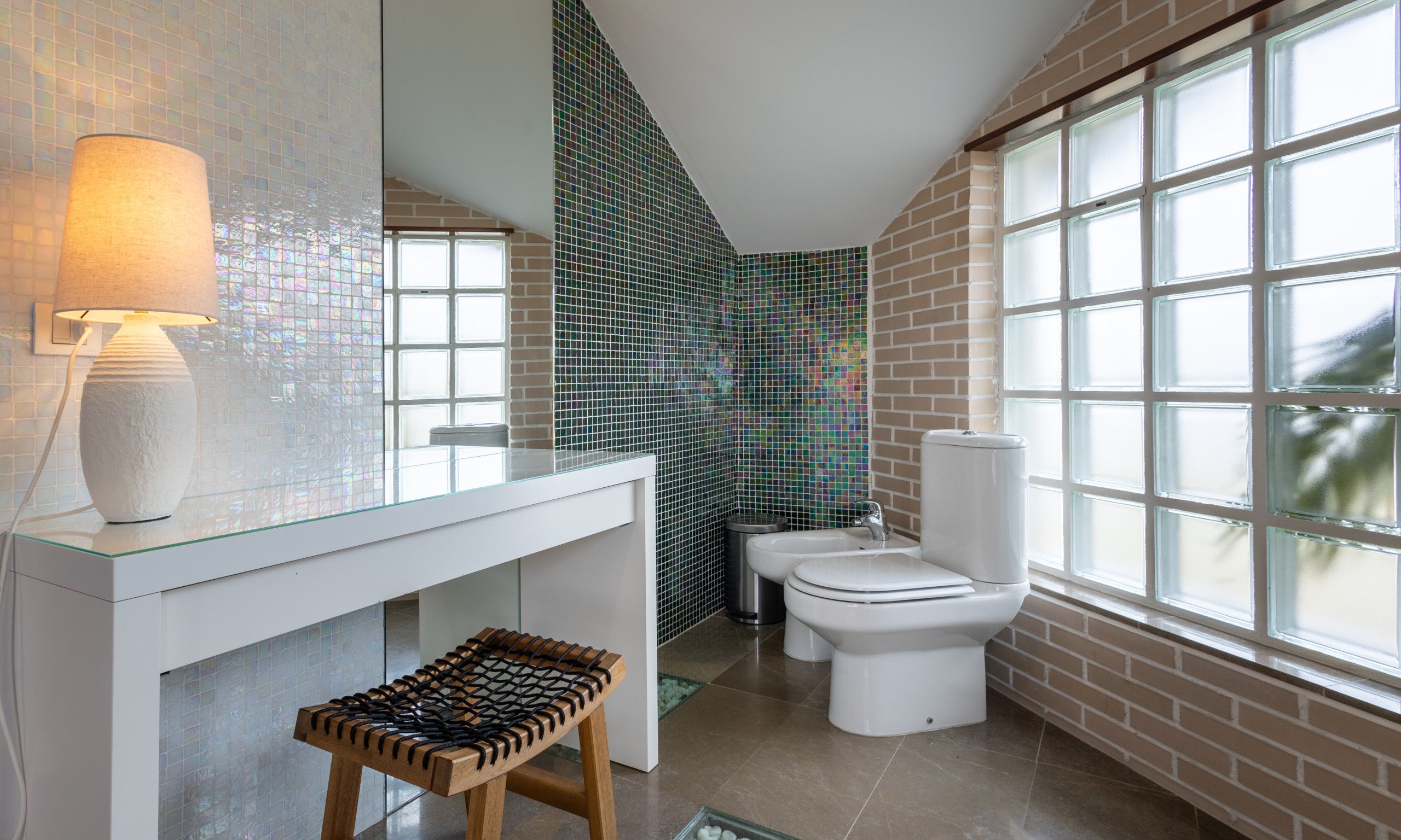 Une salle de bain moderne avec un lavabo en verre, un tabouret en bois, des toilettes blanches et un mur recouvert de carreaux.