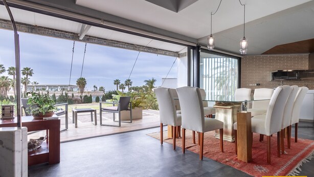 A modern dining area with a glass table and chairs, a red rug, and a view of palm trees and a building. - Ref. 494261 (Motril)