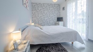 A modern bedroom with a large bed, a bedside table with a lamp, a TV on a stand, and a window with sheer curtains. - La Maiziéroise - SAVIERES (SAVIERES)