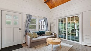 Une pièce chaleureuse avec un plafond en bois, un canapé beige agrémenté de coussins, une table basse ronde en bois et de grandes fenêtres avec des rideaux.
