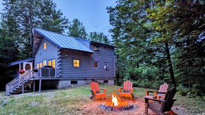 Une cabane en rondins dotée d’une terrasse et d’un foyer, le tout entouré de fauteuils Adirondack.