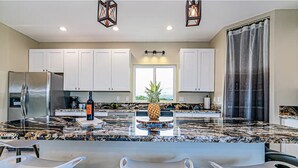 Una cocina moderna con armarios blancos, encimera de granito, refrigerador de acero inoxidable y una isla central con taburetes de bar.