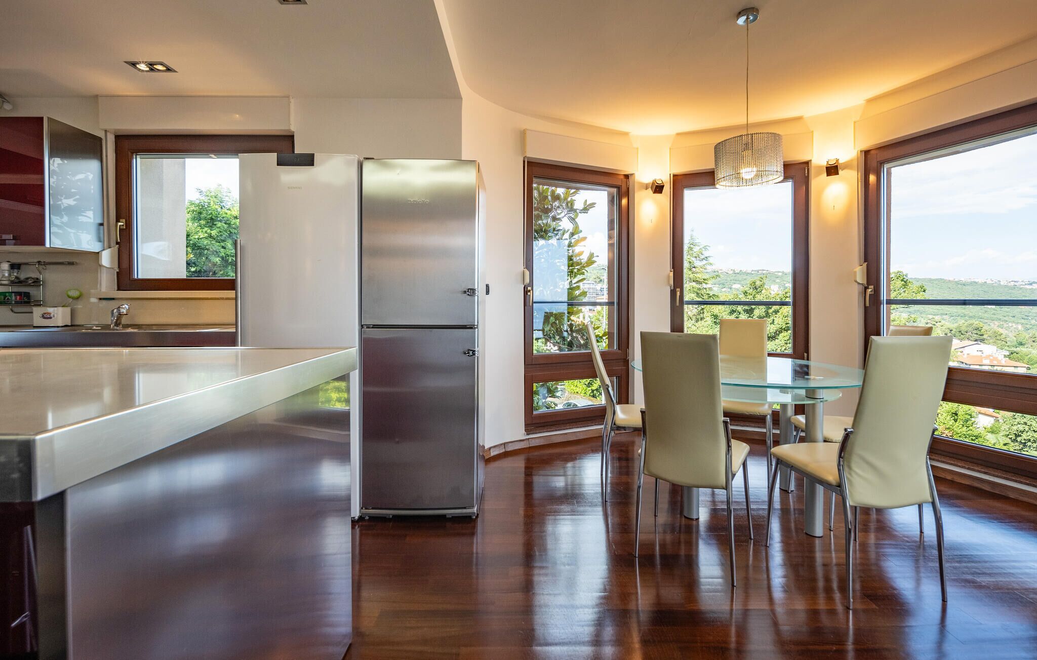 A modern kitchen with a stainless steel refrigerator, a glass dining table with four chairs, and large windows offering a view of the outdoors.