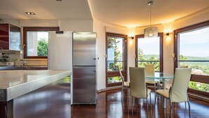 A modern kitchen with a stainless steel refrigerator, a glass dining table with four chairs, and large windows offering a view of the outdoors.