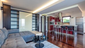 A modern living room with a grey sofa, a glass coffee table, and a patterned rug. It leads to a kitchen with red cabinets and a stainless steel refrigerator.