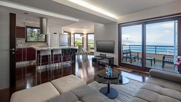 A modern living room with a sofa, a coffee table, and a TV. There is a kitchen area with a bar and a dining table. The room has large windows with a view of the sea.