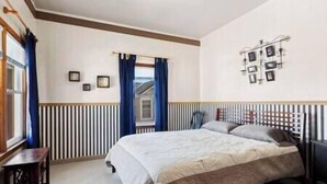 A bedroom with a bed, a desk, a window with curtains, and a wall-mounted light fixture. - Depot Suite (Elkader)