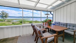 Une terrasse couverte avec une table et des chaises en bois, un canapé gris et une vue sur un lac et des collines lointaines.