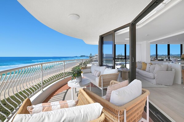 A balcony with a view of the ocean, furnished with wicker chairs and a white sofa, and a small round table with a plant.