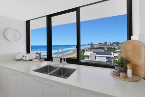 A kitchen with a large window overlooking a beach and buildings, a white clock, and a coffee maker. - Beachfront Luxury - SeaShells 3 on Albatross Ave (Mermaid Beach)
