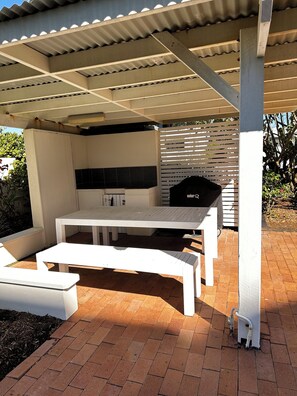 Un patio al aire libre con una parrilla, mesas blancas y piso de ladrillo.
