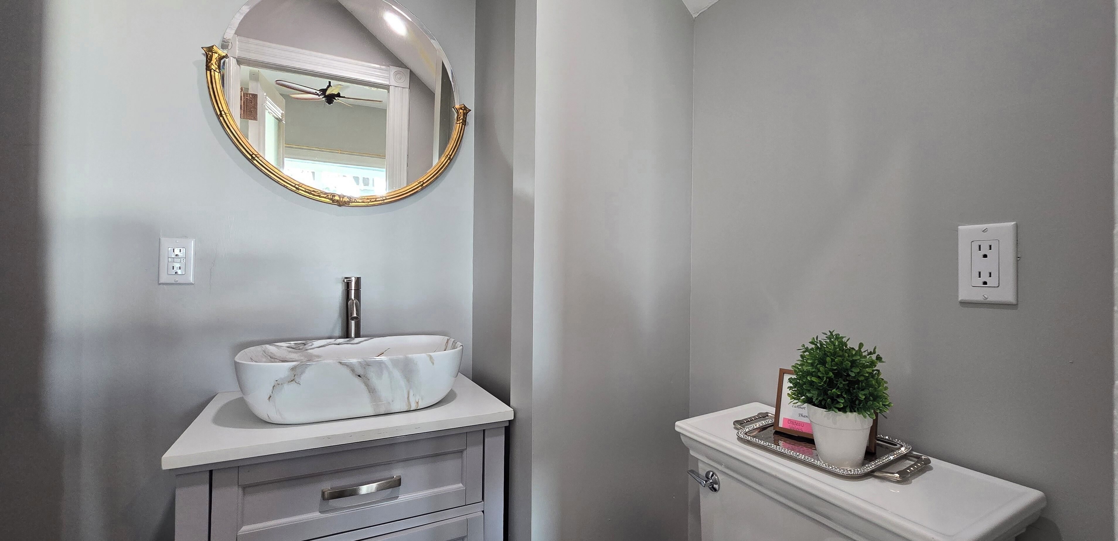 A bathroom with a marble sink, a gold-framed mirror, a potted plant, and a toilet with a tray.