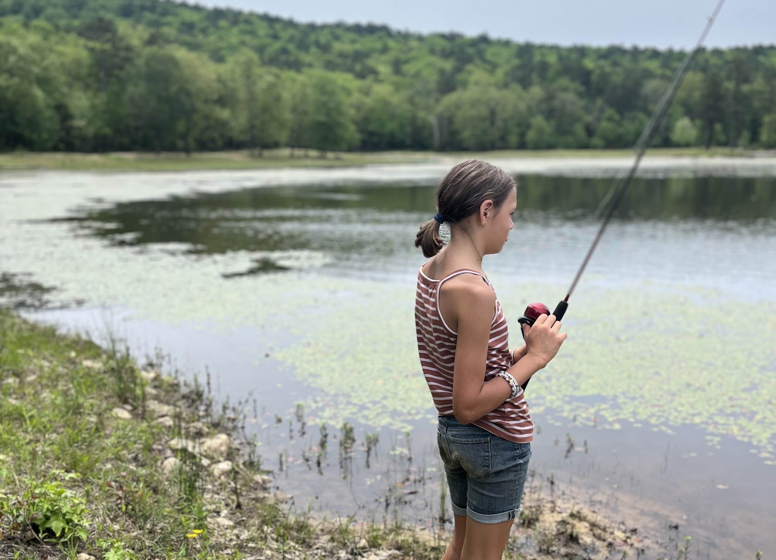 A person fishing by a calm lake with greenery in the background.