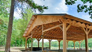 A wooden pavilion with a sloped roof, picnic tables, and a grill area, surrounded by trees in a park setting. - The Lodge at Sacred Pines (Scott County)