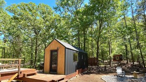 A small wooden cabin with a metal roof, surrounded by a forest. There is a wooden deck with benches and a table. - Nordic Tiny House (Scott County)