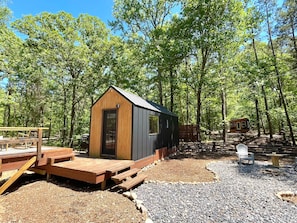 A small wooden cabin with a metal roof, surrounded by a forest. There is a wooden deck with benches and a table.
