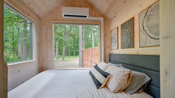 A wooden cabin bedroom with a bed, a large window, and a view of the forest. - Nordic Tiny House (Scott County)