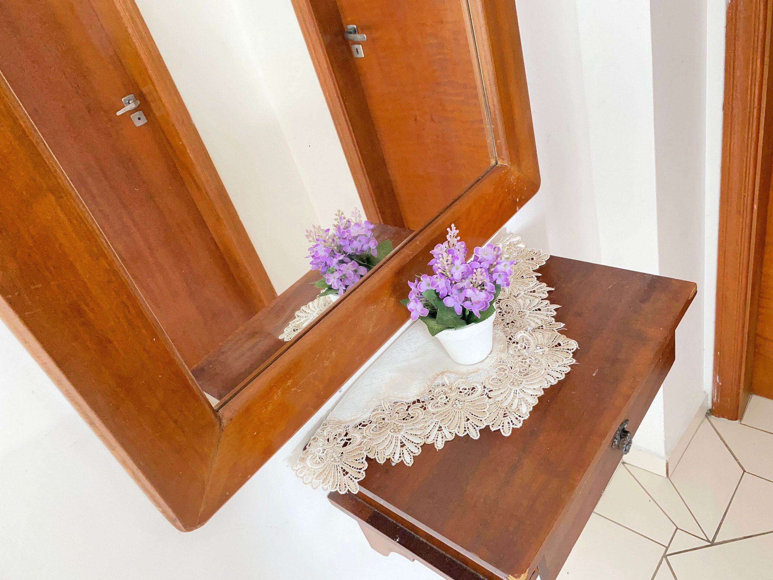 A wooden cabinet with a mirror, a lace doily, and a small white vase with purple flowers.