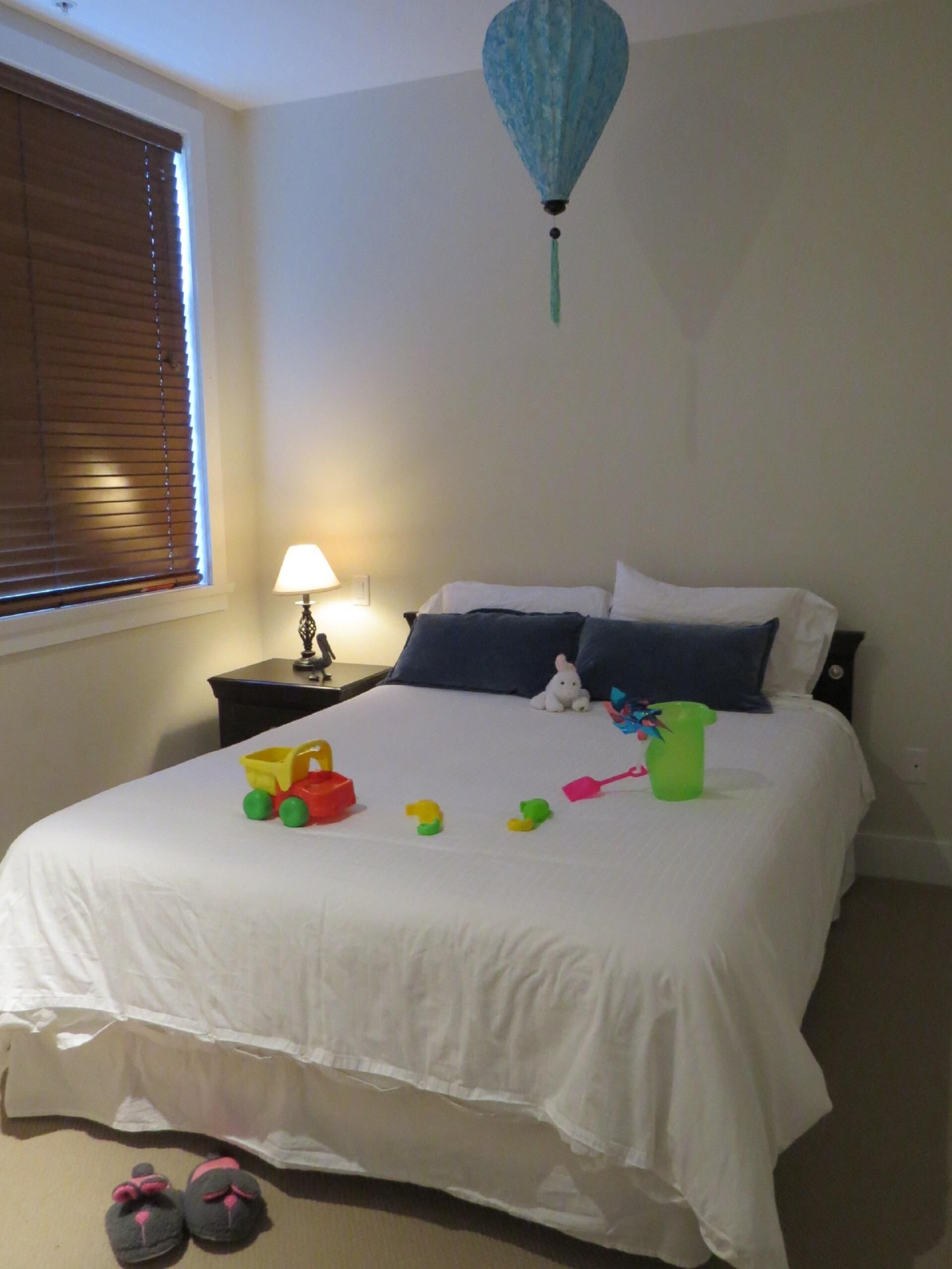 A bedroom with a bed, pillows, a nightstand, a lamp, and toys on the bed.