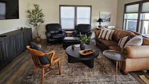 A modern living room with a dark wood floor, leather sofas, a coffee table, and a large window with blinds. - Charming New Lake House (Pierre)