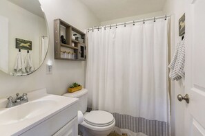 A bathroom with a white sink, toilet, and shower curtain. There is a mirror above the sink and a shelf with toiletries. - Charming 2-bedroom cottage in lovely Folsom with WiFi, AC and gorgeous views (Folsom)