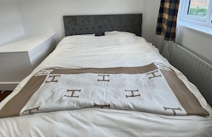 Ein ordentlich bezogenes Bett mit grauem Polsterkopfteil, einer weißen Bettdecke mit braunen Akzenten und einem Fenster mit Blick auf ein Gebäude.