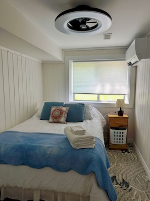 A bedroom with a bed, a window with blinds, a ceiling fan, and a nightstand with a lamp and a laundry basket.
