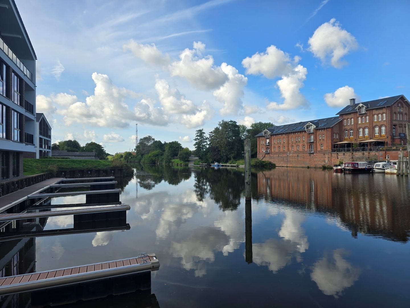 En stilla flod med förtöjda båtar, en modern byggnad till vänster och en historisk byggnad i tegel till höger.