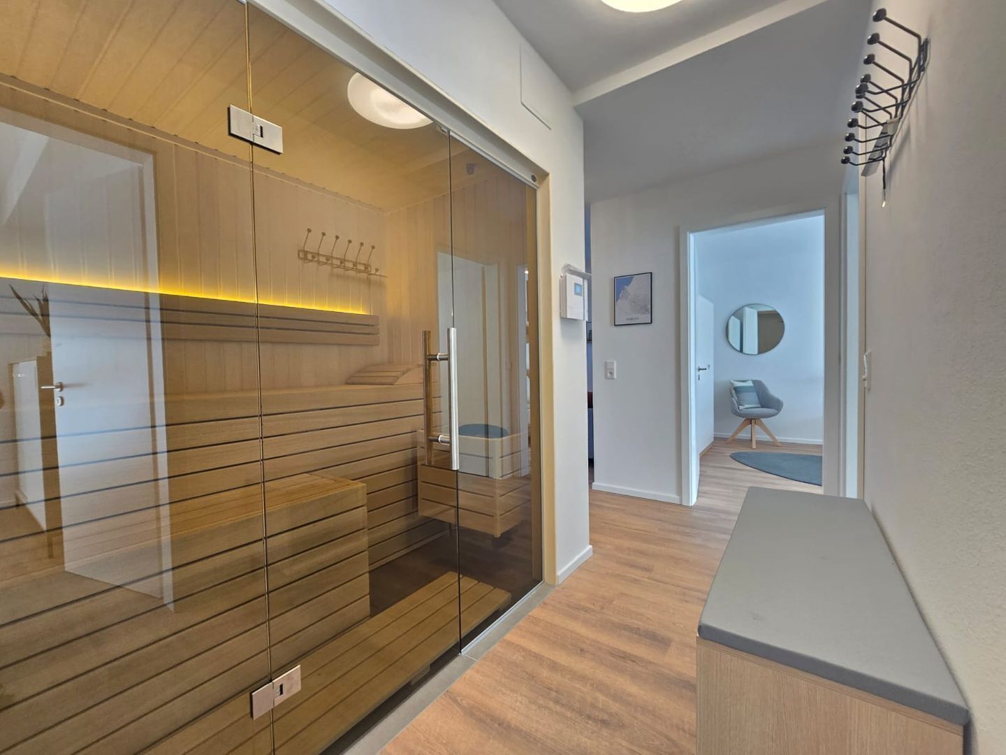 Un interior moderno con un panel de madera, una puerta de cristal y una pequeña zona de estar con un espejo redondo y una silla.