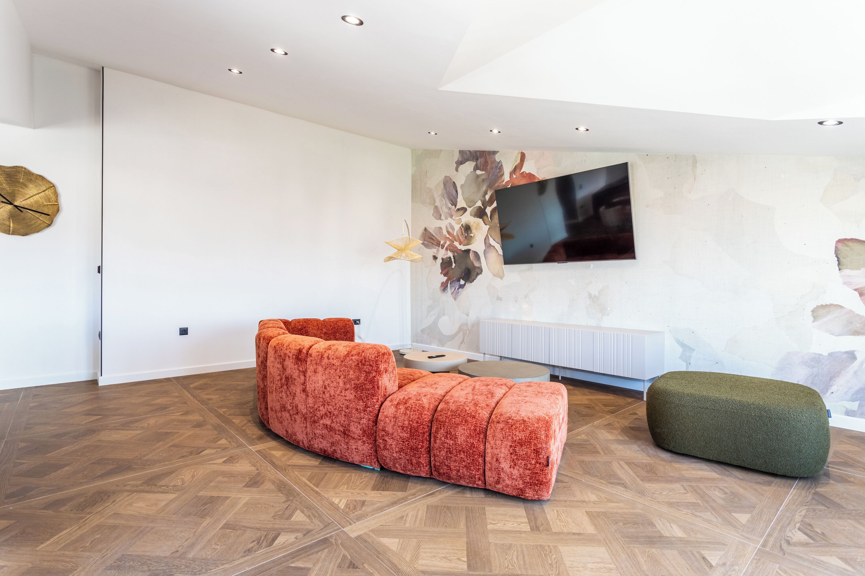 A modern living room with a sectional sofa, a green ottoman, and a flat-screen TV mounted on the wall.