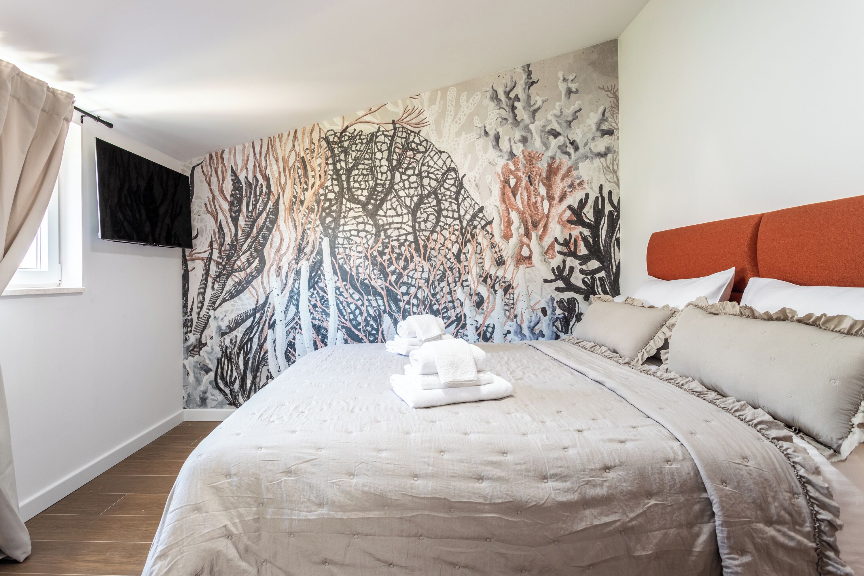 A neatly made bed with a beige bedspread and pillows, a wall mural with abstract tree-like patterns, and a television mounted on the wall.