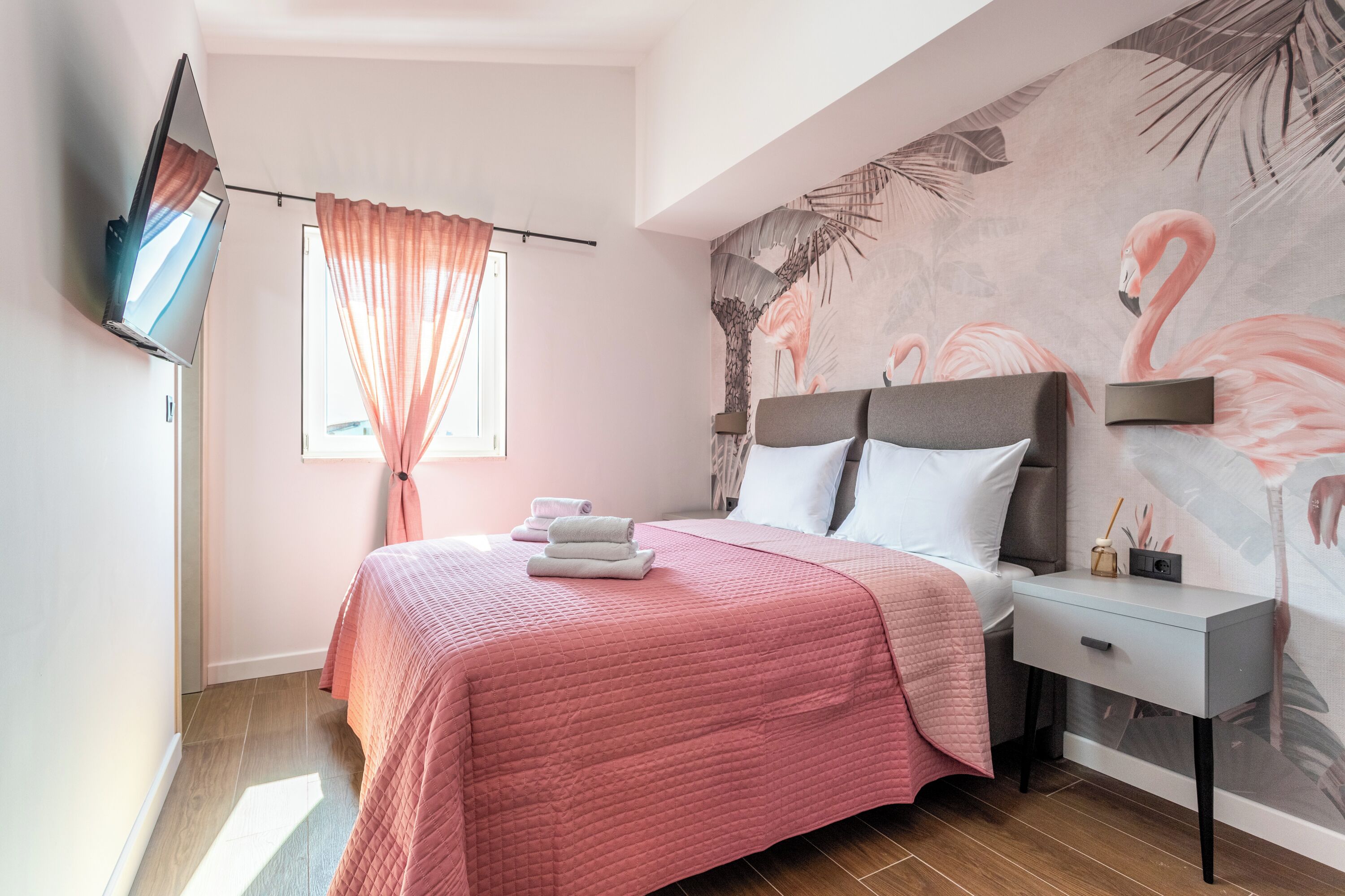 A bedroom with a bed, a nightstand, a TV mounted on the wall, and a window with curtains.