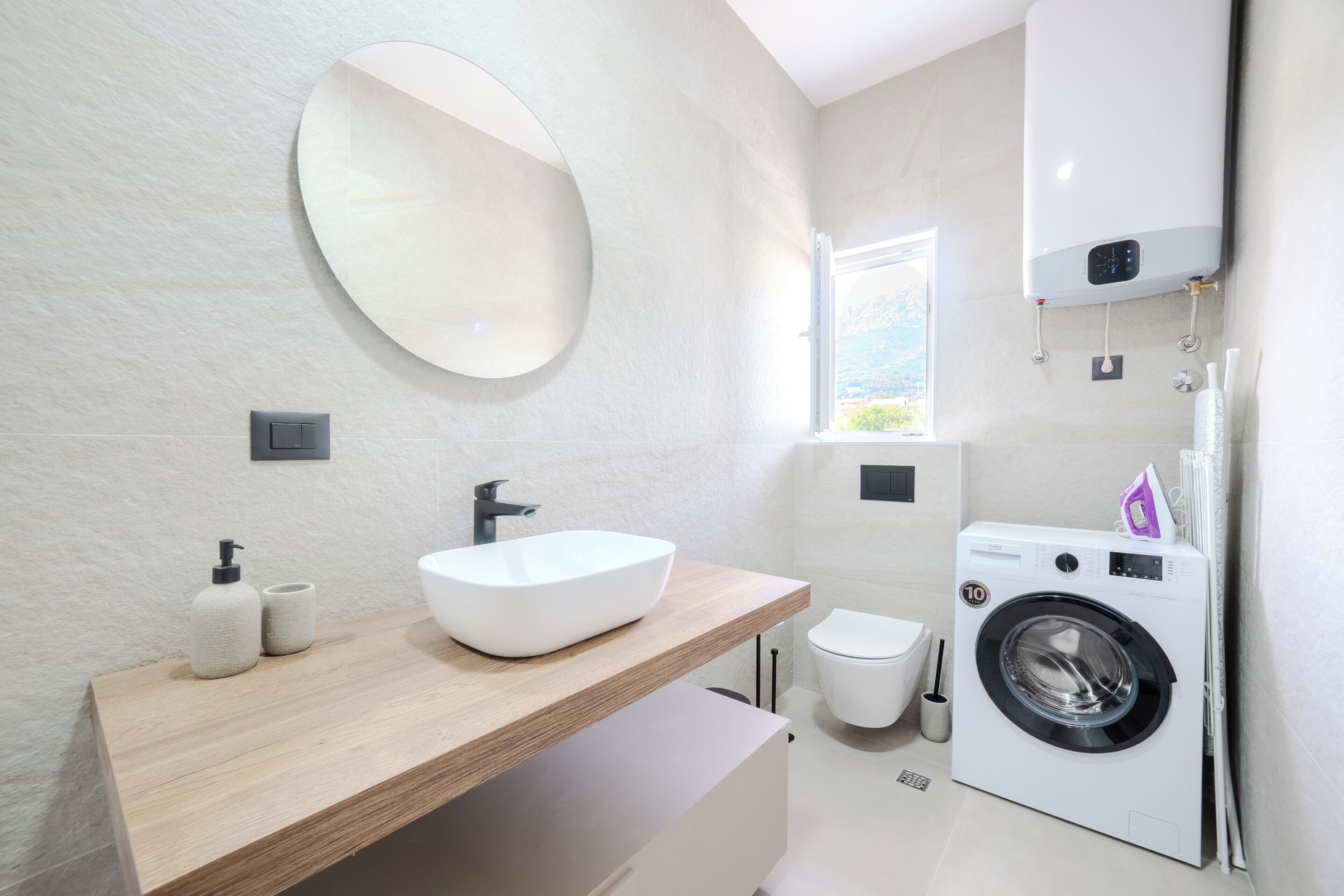 A modern bathroom with a wooden vanity, a large mirror, a white sink, a toilet, and a front-loading washing machine.