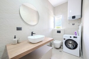 A modern bathroom with a wooden vanity, a large mirror, a white sink, a toilet, and a front-loading washing machine.