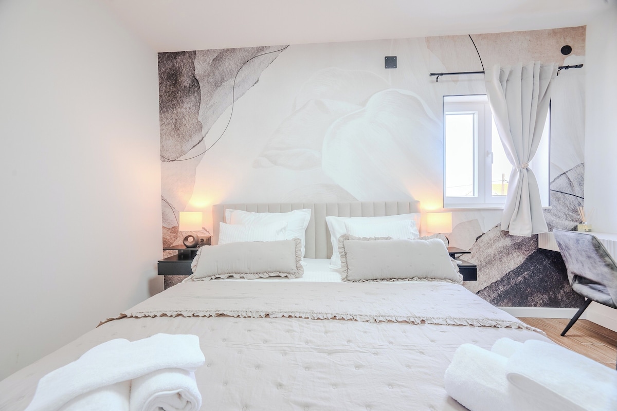 A bedroom with a large bed, two bedside tables, a window with curtains, and a wall mural.