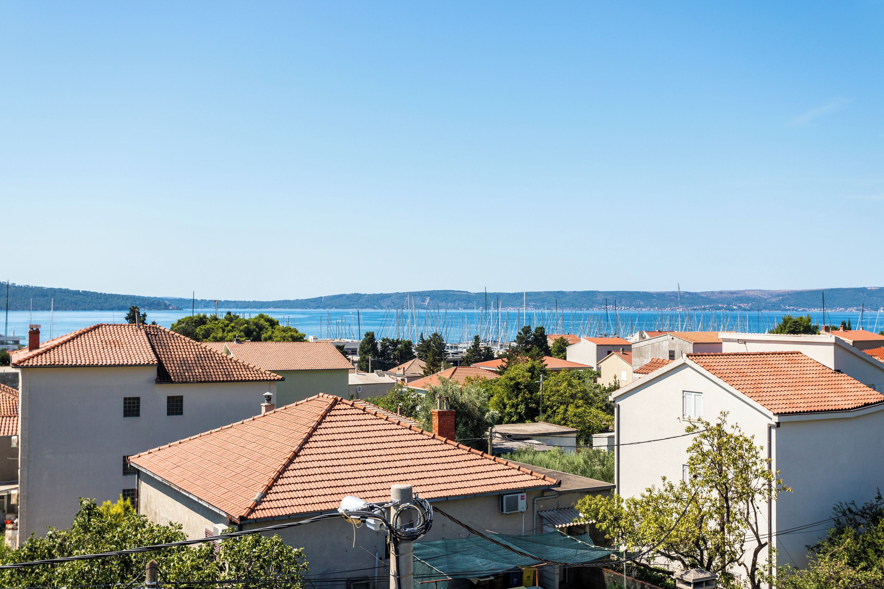 A coastal town with terracotta-roofed buildings, a marina with sailboats, and a clear blue sky.