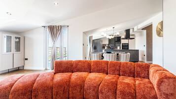 Una sala moderna con un sofá modular, una cocina al fondo y un gran ventanal con cortinas.