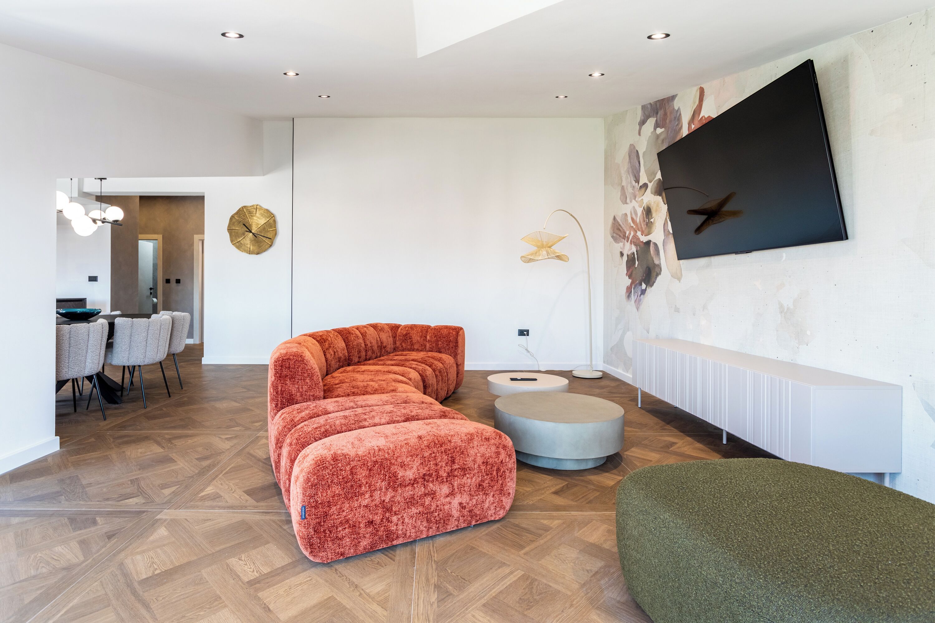 Un salon moderne avec un canapé d’angle, une table basse ronde et un pouf vert. On aperçoit à l’arrière-plan un coin repas agrémenté d’un lustre.