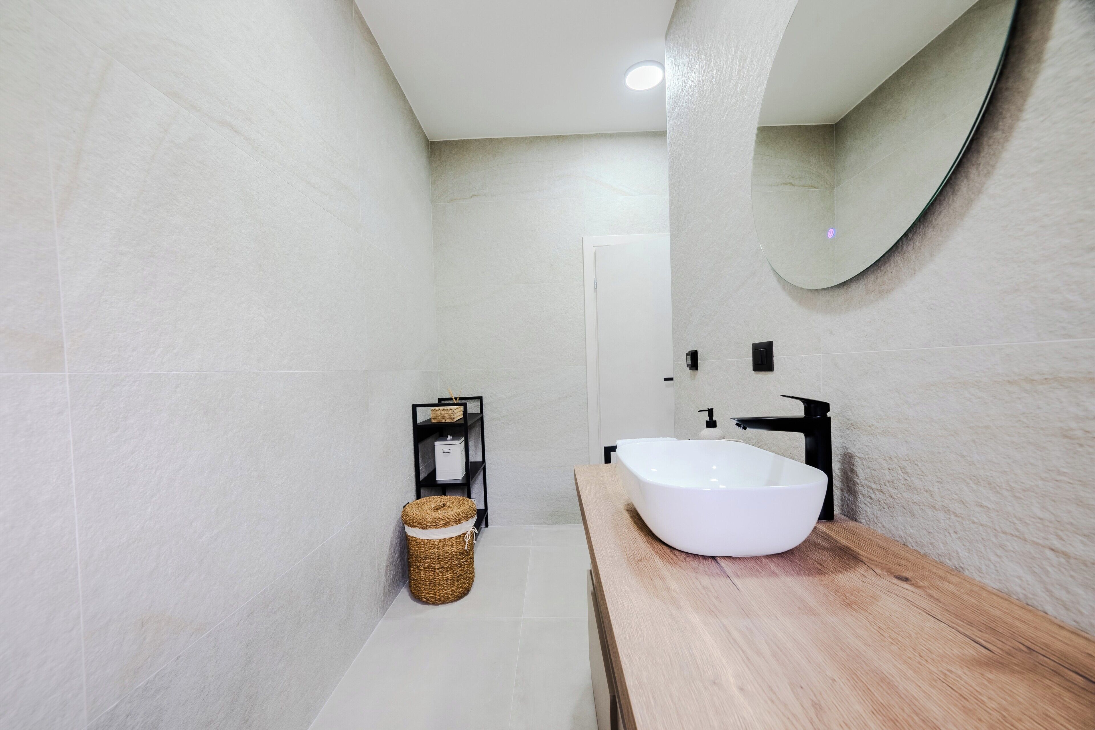 Un baño moderno con un lavamanos grande blanco, un mueble de tocador de madera, un estante negro con artículos de aseo y un espejo redondo.