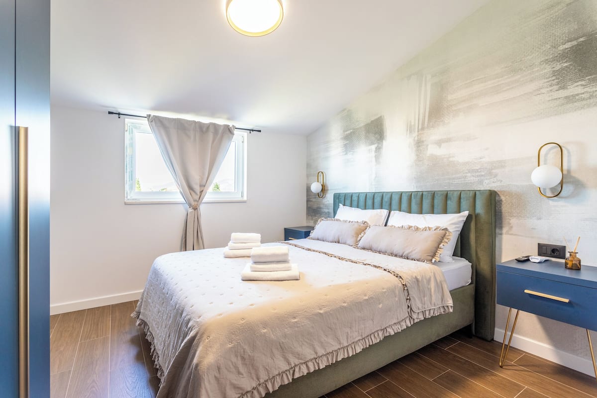 A modern bedroom with a large bed, a nightstand, a wall-mounted lamp, and a window with curtains.