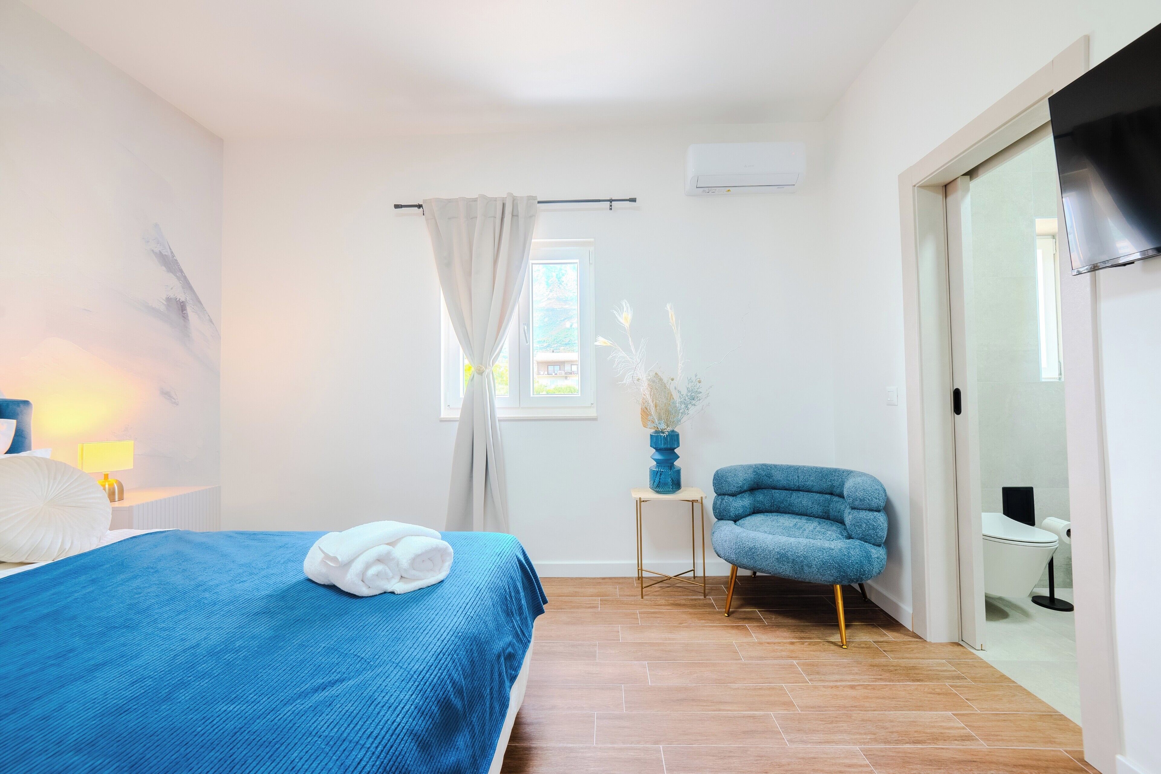 Habitación de hotel con cama, una silla, una mesita, ventana con cortinas y baño visible a través de una puerta abierta.