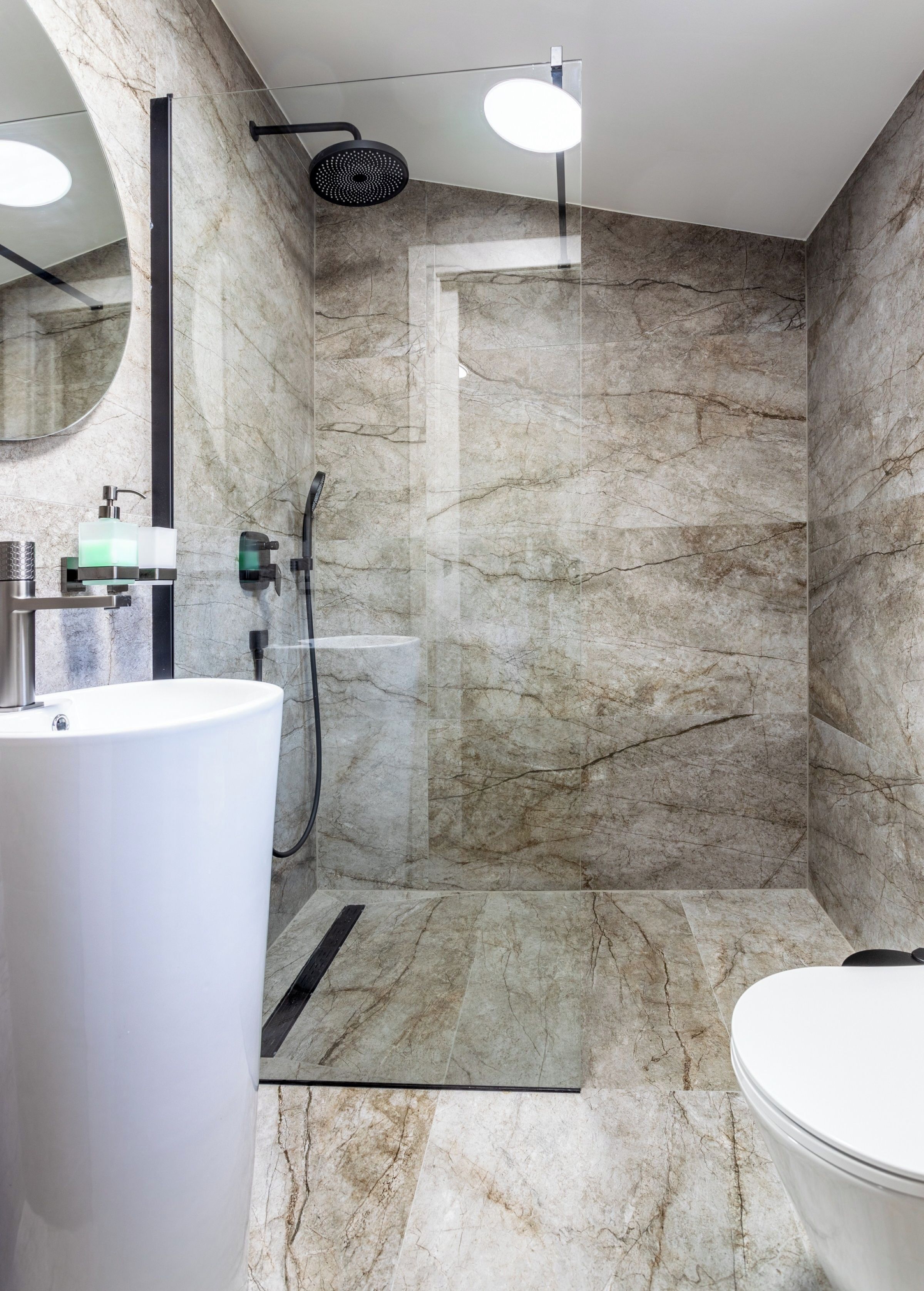 A modern bathroom with a marble wall, a large white sink, a toilet, and a shower area with a glass door.