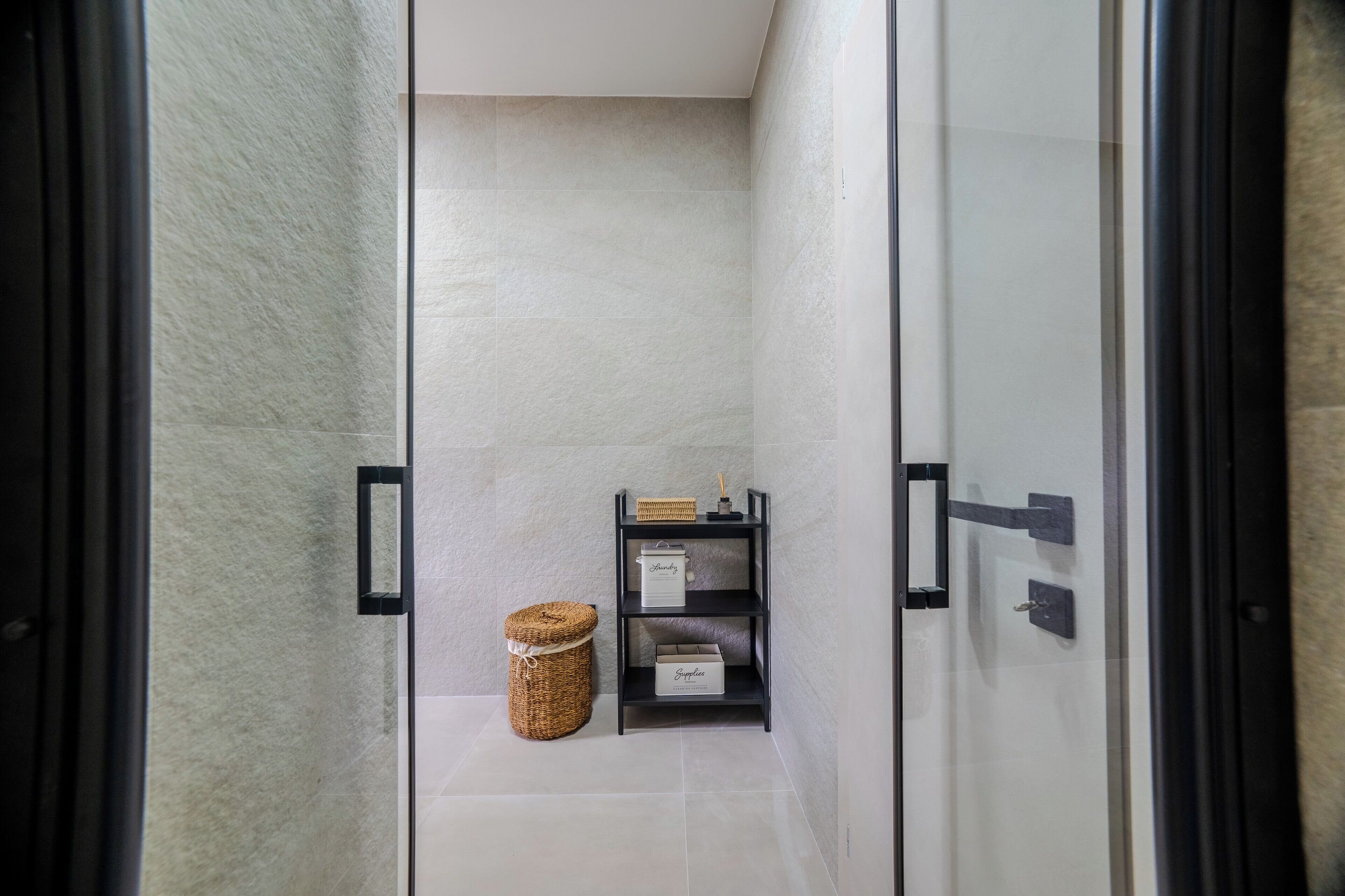 A narrow hallway with a glass door leading to a room. There is a wicker basket and a shelf with items on it.