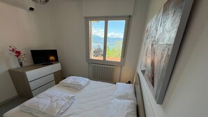 A neatly made bed with white linens, a bedside table with a flat-screen TV, a window with a view of greenery and mountains, and a painting on the wall. - Marenco apartment in Stresa with lake view (Stresa)