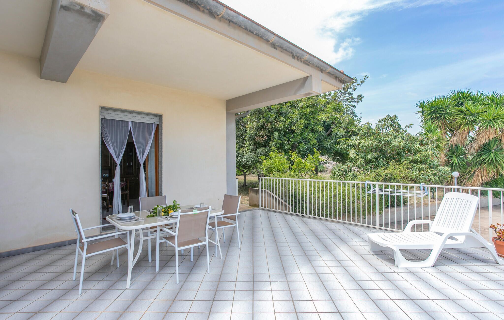 A covered outdoor patio with a table and chairs, a white railing, and a view of greenery.