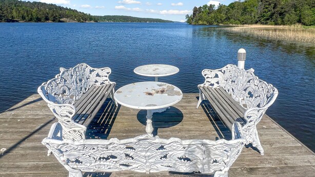 Een houten steiger met sierlijke witte metalen stoelen en een kleine ronde tafel, met uitzicht op een rustig meer met verre bomen en heuvels.