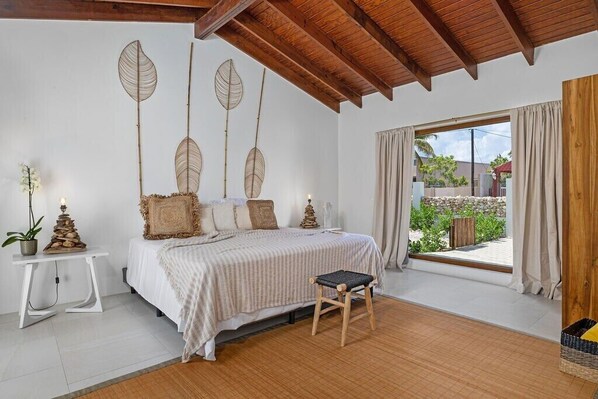 A bedroom with a bed, a small stool, a nightstand, and decorative leaf wall art. - Buddha House 5/5 Palapa Suite Pool in Palm Beach (Noord)