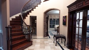 Una imponente escalera con barandillas decorativas que conduce a un rellano en el segundo piso. Se observa un pasillo con piso de cuadros y una puerta con paneles de vidrio.