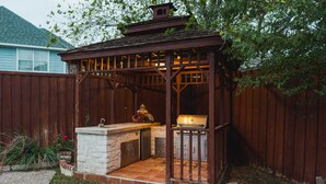 Una pérgola de madera con encimera de piedra y parrilla, rodeada por una valla de madera.