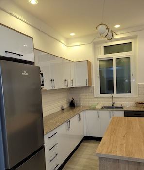 Een moderne keuken met witte kasten, een roestvrijstalen koelkast, een granieten aanrechtblad en een raam met binnengebroken glas.